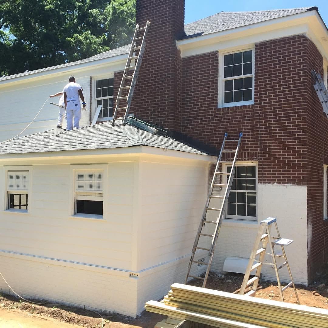 A painter on a ladder working along the upper wall of a house, ladders and equipment set up below.