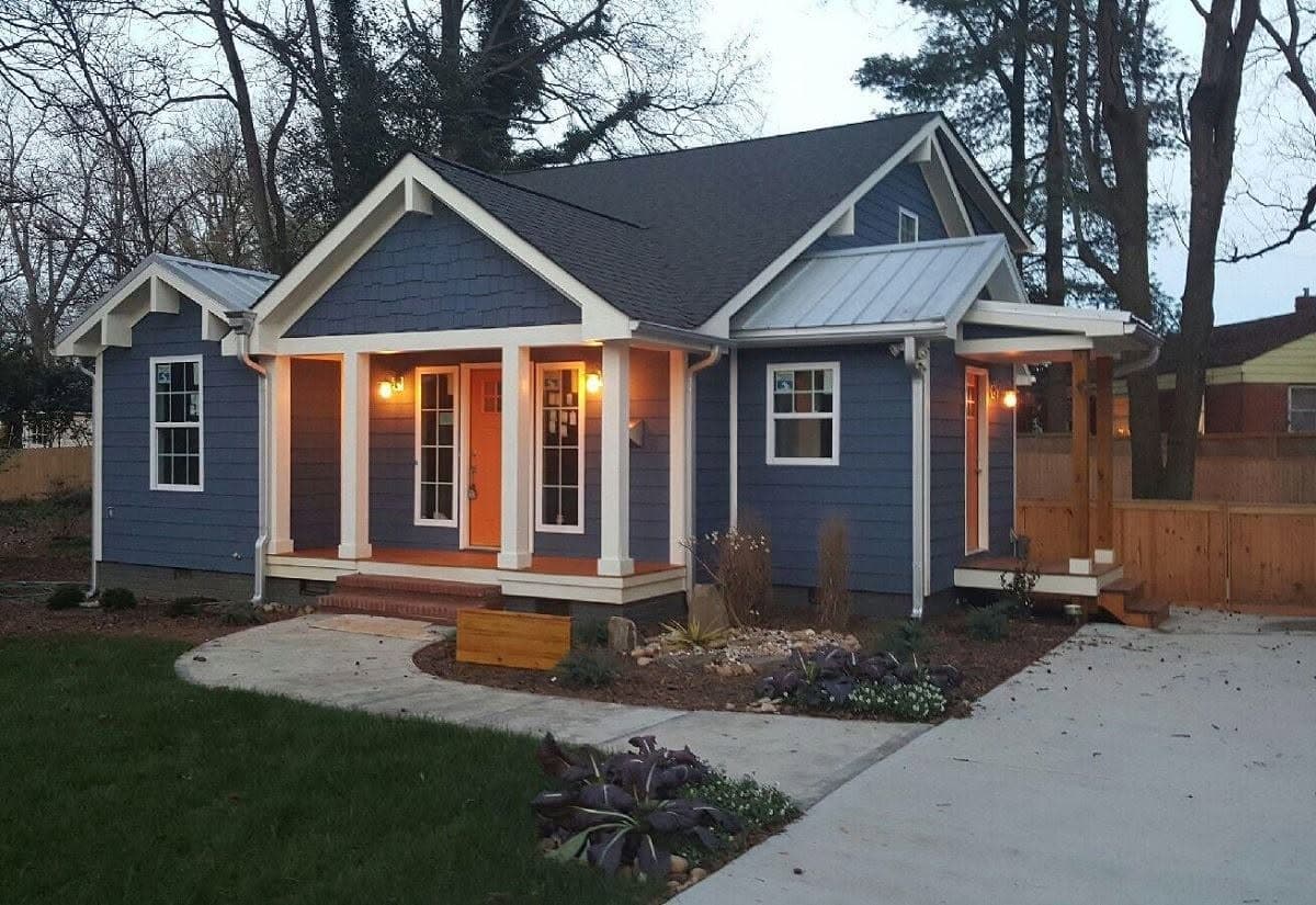 The same bungalow finished at dusk, painted blue-grey with cream trim, a warm-toned front door and the porch lights on.