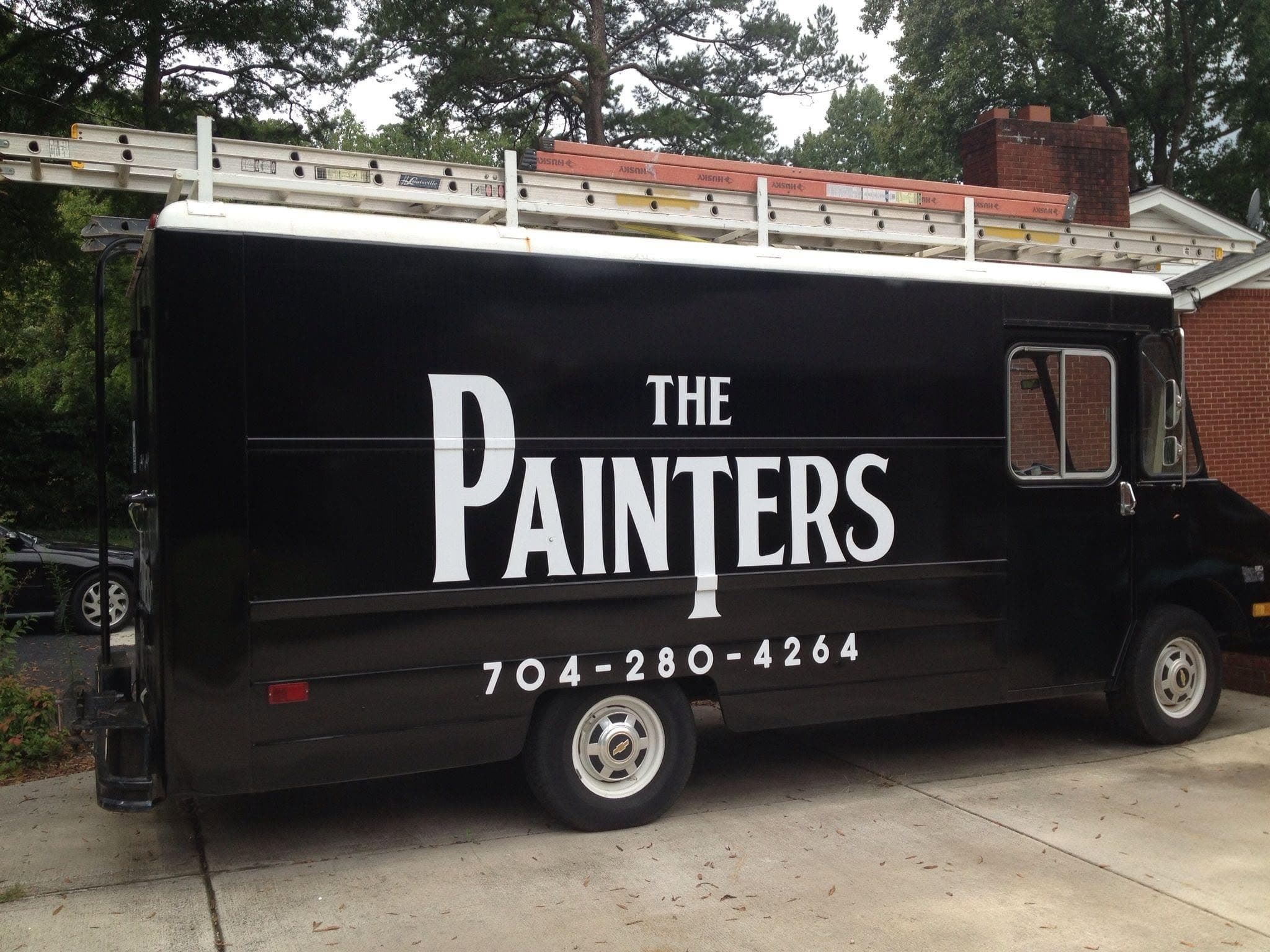 The Painters black box truck, sign-written with the company name and phone number, with ladders racked on top.